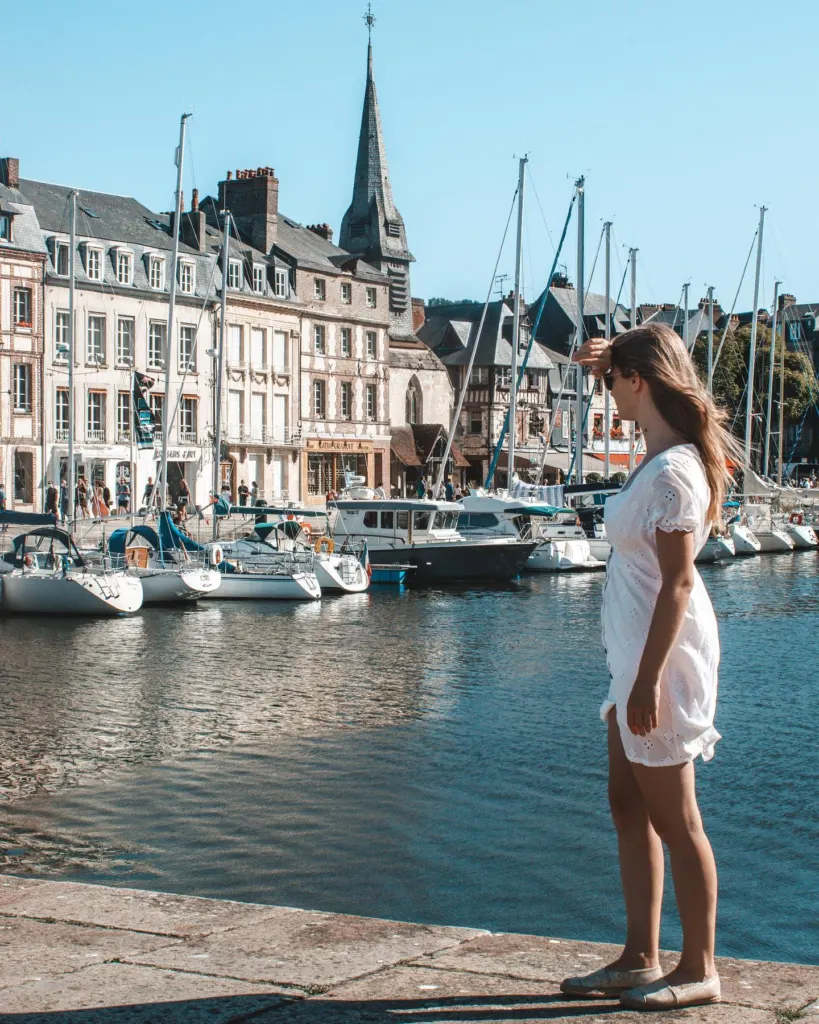 Woman by the harbour of a Normandy port town with tall old houses and a church spire