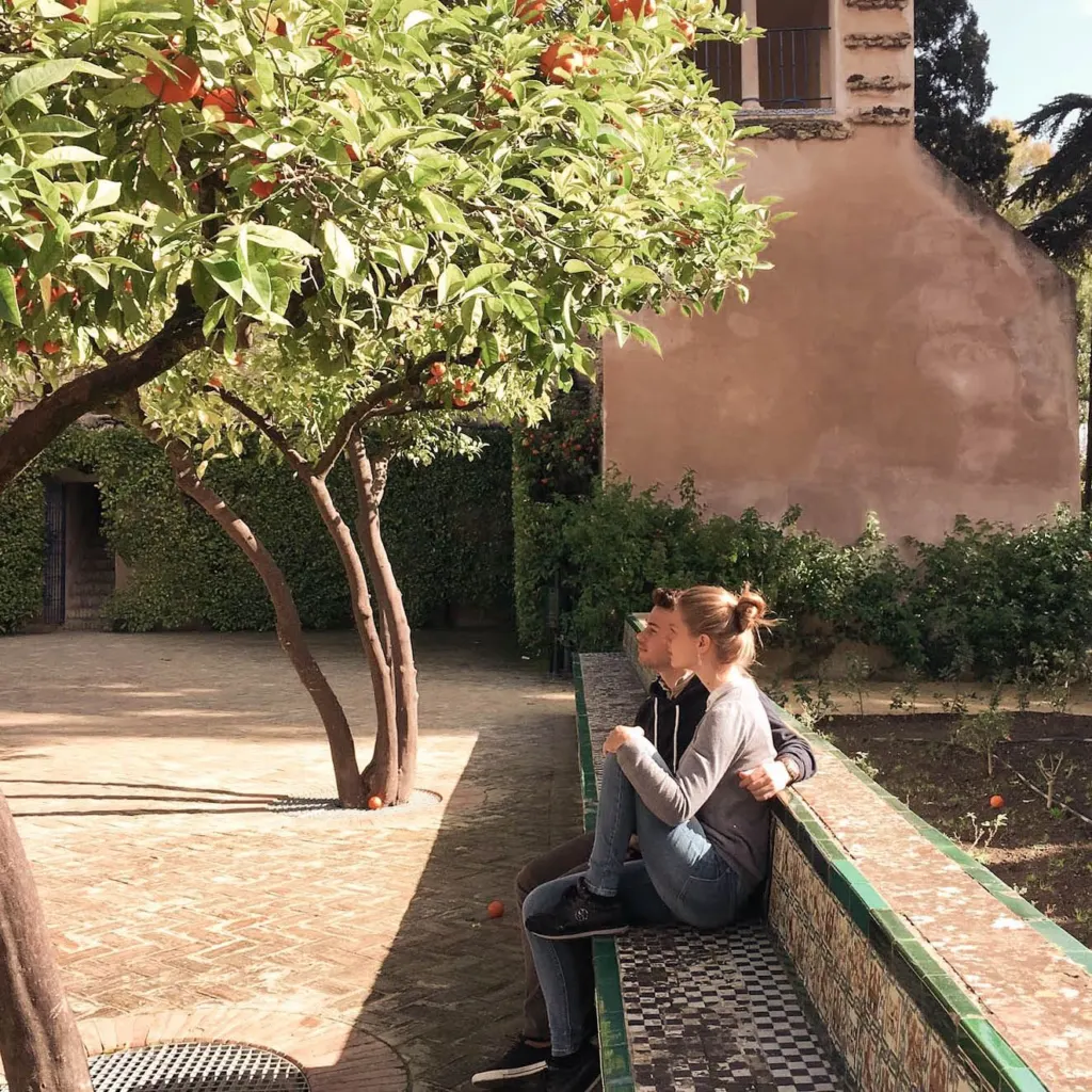 Two people sitting on a tiled bench under an orange tree in a Seville garden